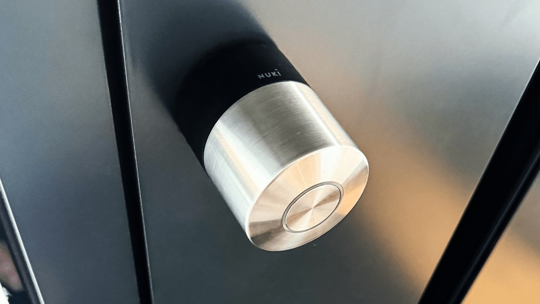 A Nuki Smart Lock, featuring a black body and a brushed silver cap, is mounted on a sleek, dark grey aluminum pivot door manufactured by AVS. The lock is seen from a slightly elevated, close-up perspective, highlighting its metallic sheen.