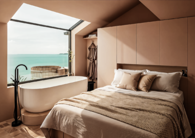 A modern bedroom with neutral tones featuring a freestanding white bathtub on a raised platform next to a large window with a view of the ocean and a wooden hot tub on a deck. A king-size bed with beige linens is also visible.