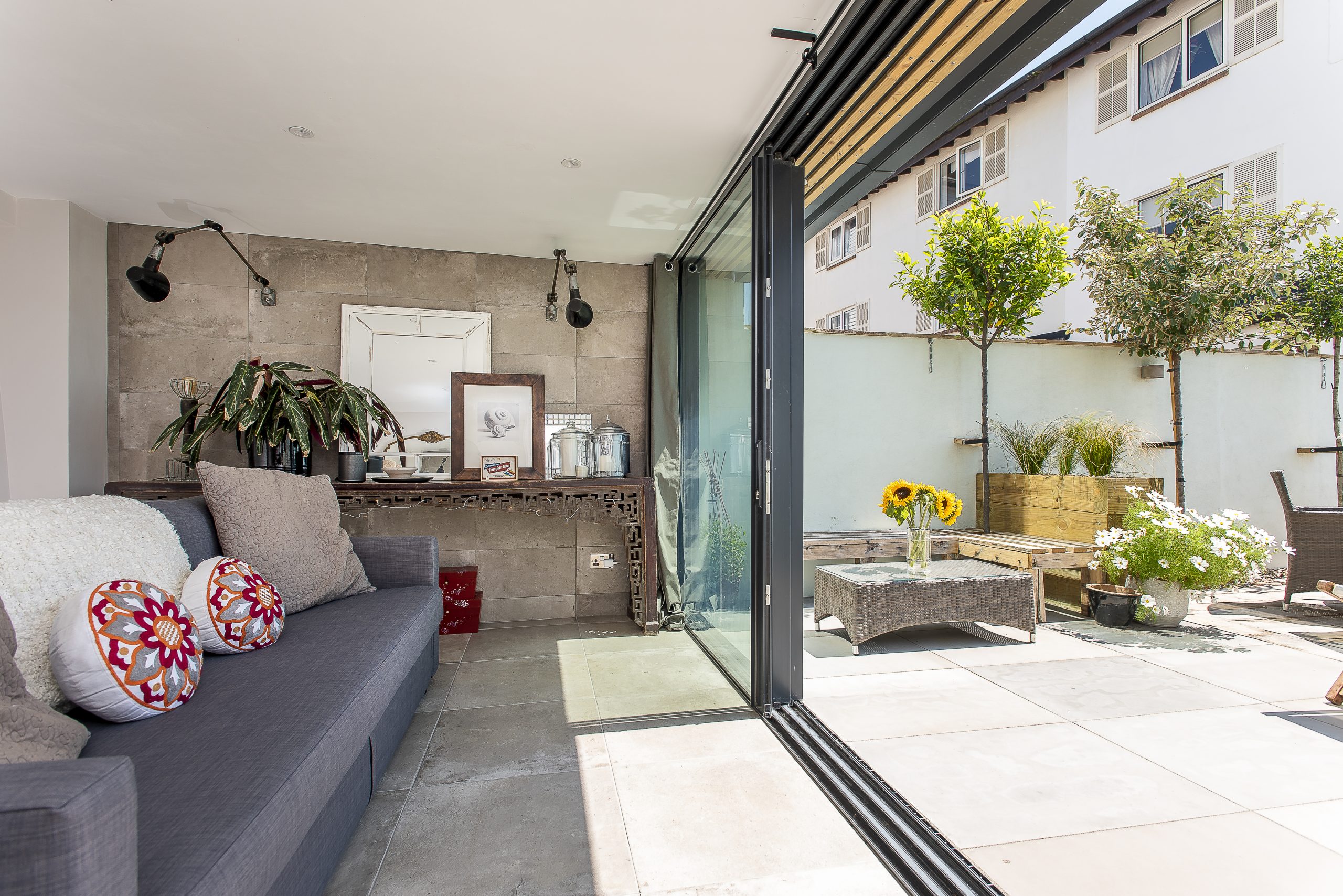 A modern bedroom with neutral tones featuring a freestanding white bathtub on a raised platform next to a large window with a view of the ocean and a wooden hot tub on a deck. A king-size bed with beige linens is also visible.
